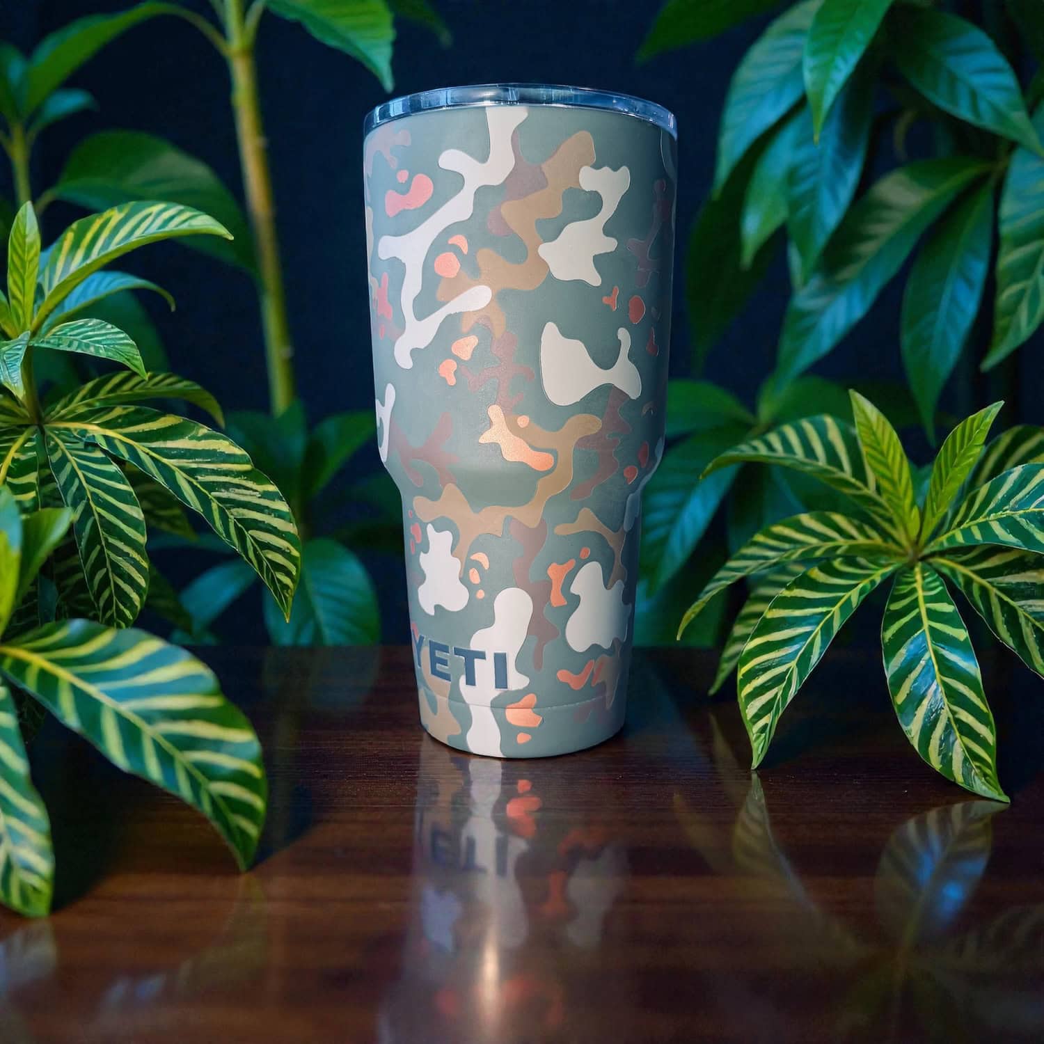 Camouflage-patterned tumbler with YETI logo surrounded by green tropical plants.